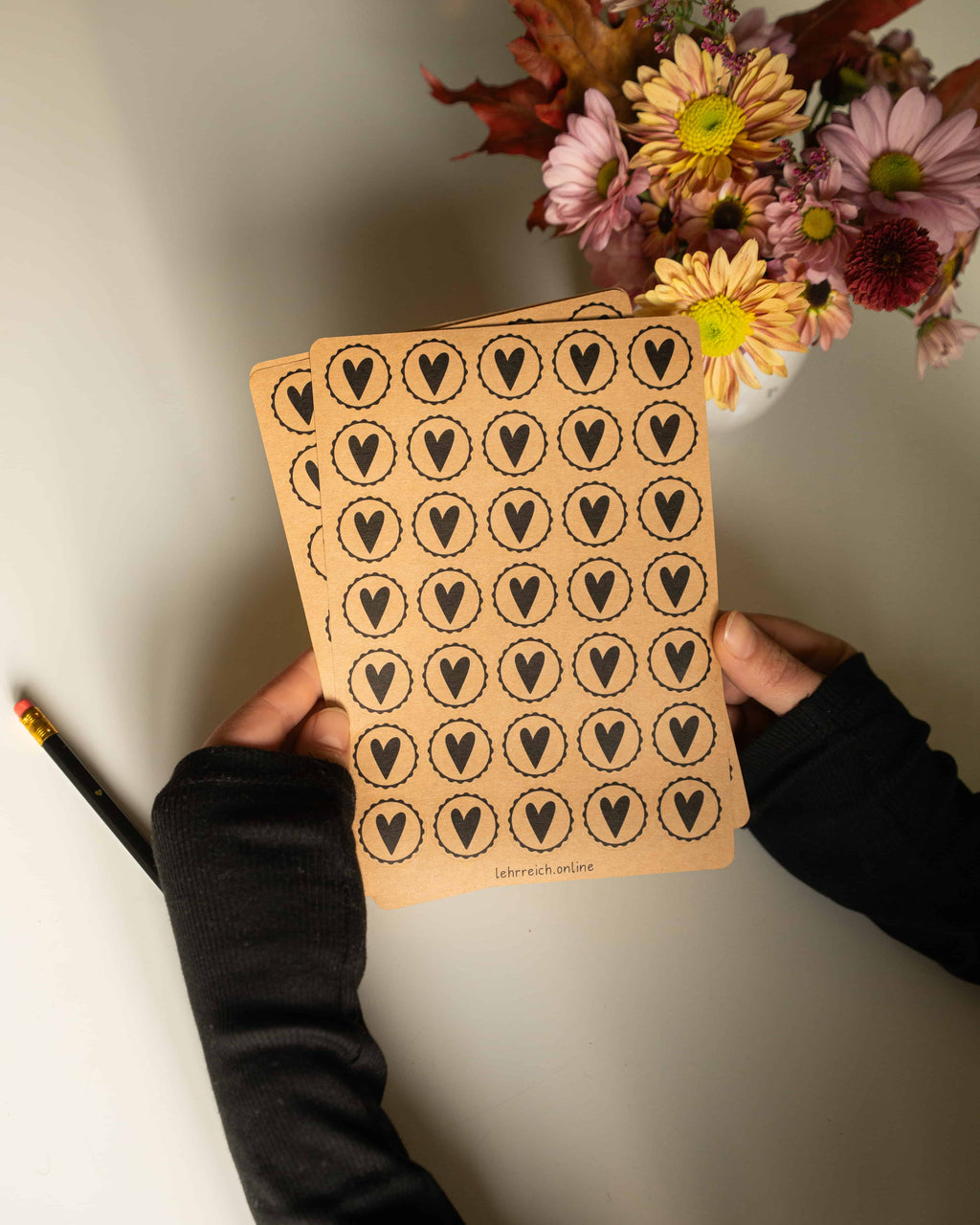 Zwei Hände halten einen Kraftpapier-Stickerbogen mit runden Herzstickern in schwarzem Druck und gewellter Kontur; warmes Licht und Blumen im Hintergrund sorgen für eine gemütliche, handgemachte Atmosphäre.